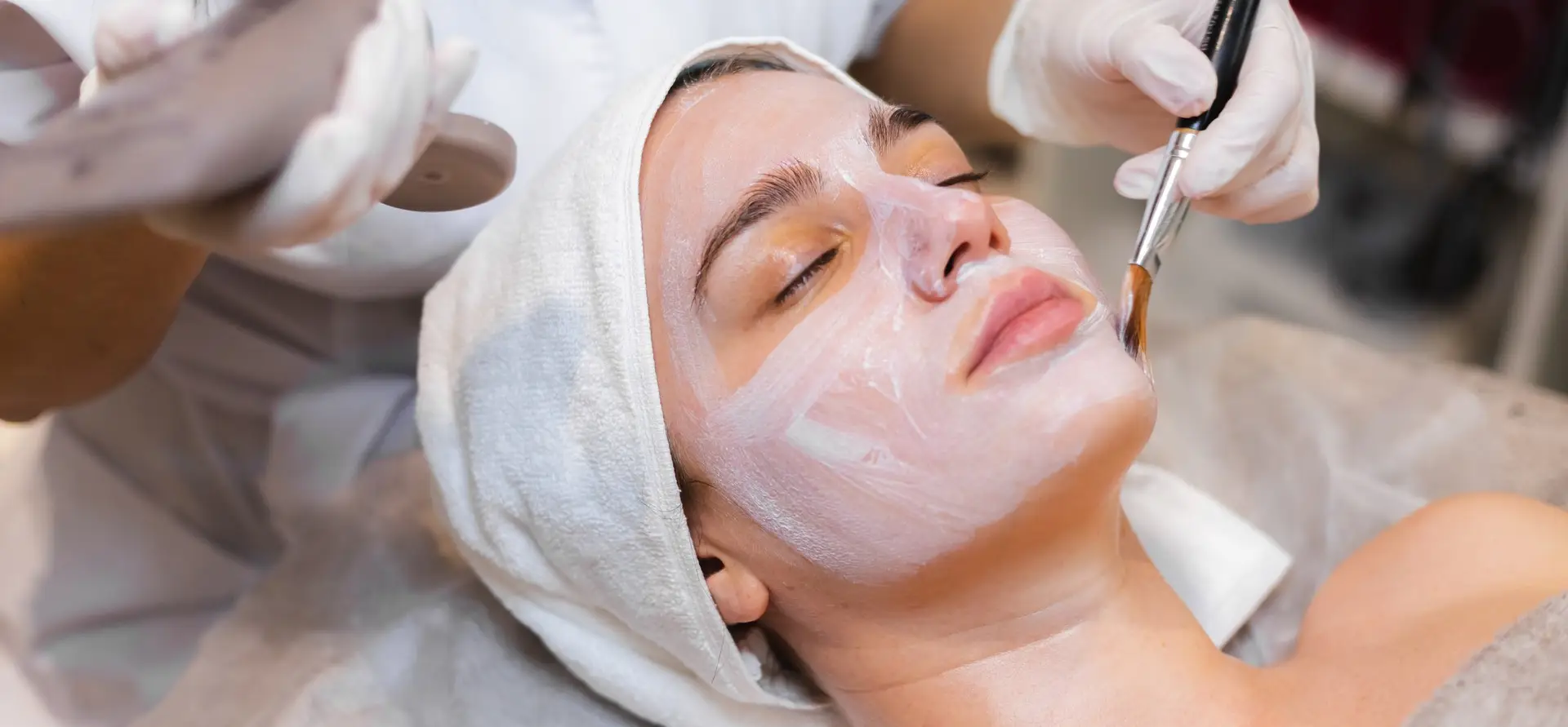 A person receiving a facial treatment, with hands in gloves applying product using a brush, surrounded by a cozy spa setting.